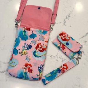 May include: A pink crossbody bag with a matching small wallet and wrist strap, all featuring a repeating pattern of a red-haired mermaid, yellow fish, and blue coral. The bag has a pink interior lining, a snap closure, and a pink adjustable strap.