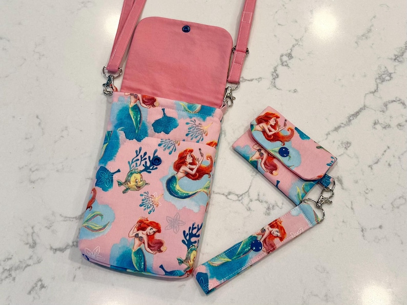 May include: A pink crossbody bag with a matching small wallet and wrist strap, all featuring a repeating pattern of a red-haired mermaid, yellow fish, and blue coral. The bag has a pink interior lining, a snap closure, and a pink adjustable strap.