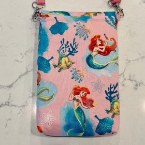 May include: A rectangular fabric crossbody bag featuring a light pink background with a repeating pattern of the mermaid character Ariel, a yellow fish, blue coral, and white floral outlines. The bag includes a pink strap attached with silver hardware.