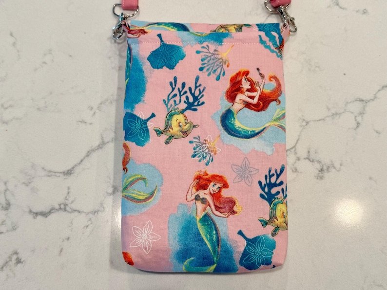 May include: A rectangular fabric crossbody bag featuring a light pink background with a repeating pattern of the mermaid character Ariel, a yellow fish, blue coral, and white floral outlines. The bag includes a pink strap attached with silver hardware.