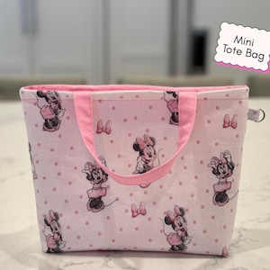 May include: Pink mini tote bag with a repeating pattern of Minnie Mouse and bows on a dotted background. The bag has pink handles and trim, and the text "Mini Tote Bag" is on a pink label.