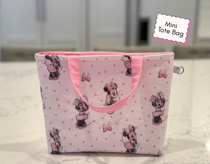 May include: Pink mini tote bag with a repeating pattern of Minnie Mouse and bows on a dotted background. The bag has pink handles and trim, and the text "Mini Tote Bag" is on a pink label.