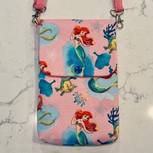 May include: A pink crossbody bag featuring a repeating pattern of a red-haired mermaid and a yellow fish among blue coral and white flower outlines. The bag has a fold-over flap secured with a dark blue snap button and pink fabric straps.