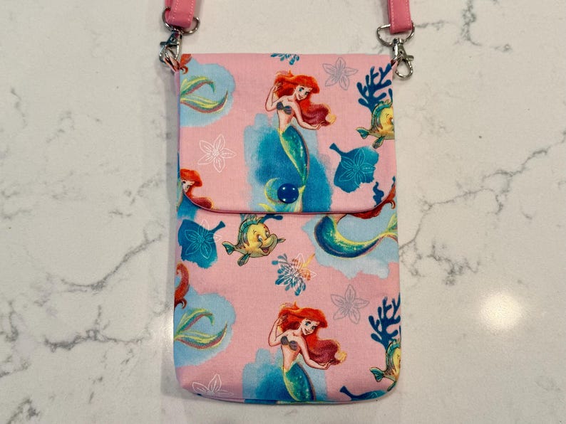 May include: A pink crossbody bag featuring a repeating pattern of a red-haired mermaid and a yellow fish among blue coral and white flower outlines. The bag has a fold-over flap secured with a dark blue snap button and pink fabric straps.