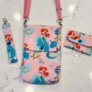 May include: A pink crossbody bag, a matching wristlet strap, and a small rectangular wallet, all featuring a repeating pattern of a red-haired mermaid, yellow fish, and blue coral on a light pink background with dark blue snap button closures.