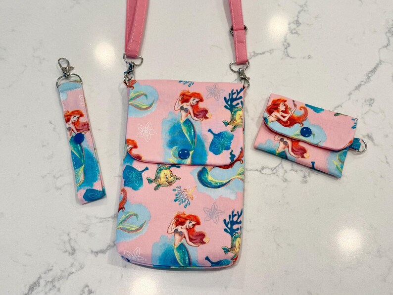 May include: A pink crossbody bag, a matching wristlet strap, and a small rectangular wallet, all featuring a repeating pattern of a red-haired mermaid, yellow fish, and blue coral on a light pink background with dark blue snap button closures.