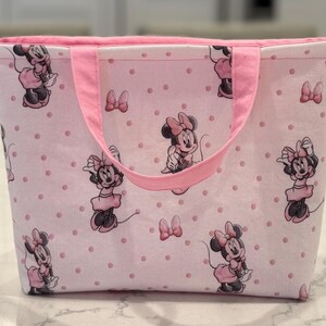 May include: Pink tote bag featuring a pattern of Minnie Mouse illustrations and pink polka dots. The bag has pink handles and a pink interior. A small, silver-colored ring is attached to the side.