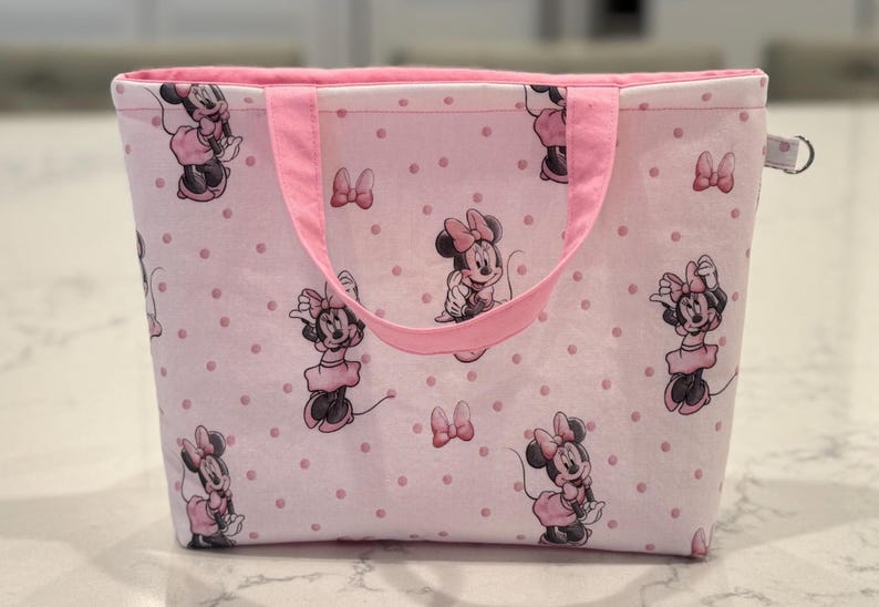 May include: Pink tote bag featuring a pattern of Minnie Mouse illustrations and pink polka dots. The bag has pink handles and a pink interior. A small, silver-colored ring is attached to the side.