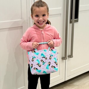 May include: A small tote bag with a mermaid pattern in shades of purple, teal, and black. The bag has teal handles and a rectangular shape. The bag is held by a person wearing a pink zip-up hoodie and black pants.