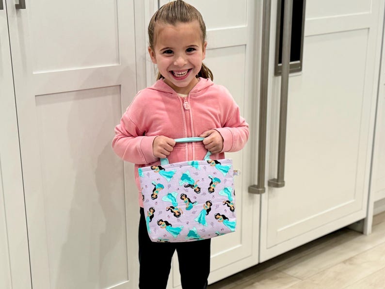 May include: A small tote bag with a mermaid pattern in shades of purple, teal, and black. The bag has teal handles and a rectangular shape. The bag is held by a person wearing a pink zip-up hoodie and black pants.