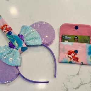 May include: A set featuring a purple sequined headband with a light blue sequined bow, a matching fabric keychain strap with an Ariel graphic, and a small pink fabric wallet with an Ariel print containing a Disney Premier card.