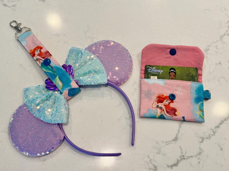 May include: A set featuring a purple sequined headband with a light blue sequined bow, a matching fabric keychain strap with an Ariel graphic, and a small pink fabric wallet with an Ariel print containing a Disney Premier card.