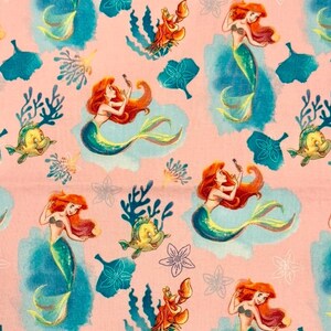 May include: A light pink fabric featuring a repeating pattern of a red-haired mermaid with a green tail, a yellow fish, a red crab, and blue watercolor-style coral and floral shapes. The design includes various aquatic elements in blue.