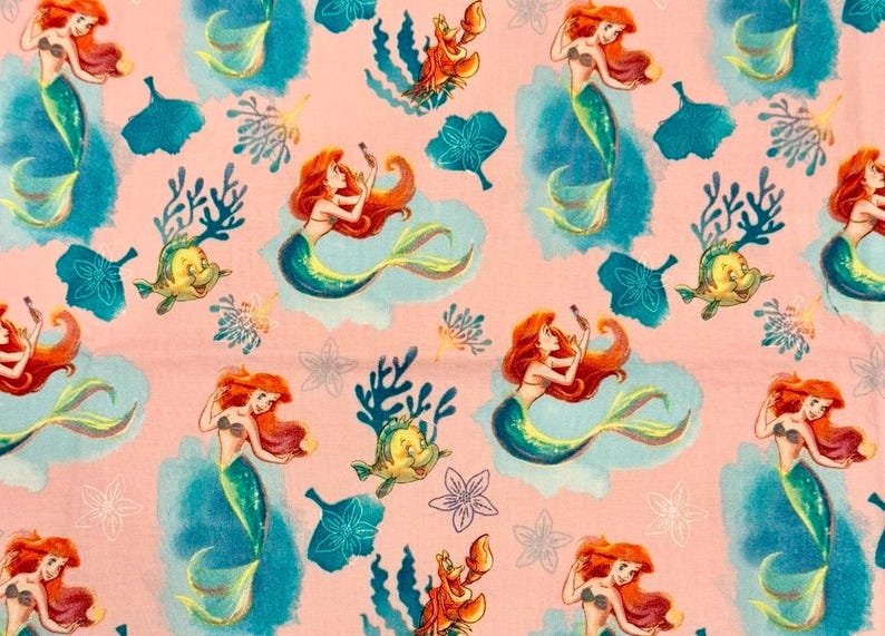 May include: A light pink fabric featuring a repeating pattern of a red-haired mermaid with a green tail, a yellow fish, a red crab, and blue watercolor-style coral and floral shapes. The design includes various aquatic elements in blue.