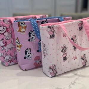 May include: Three fabric tote bags with different patterns. One bag features pink cats, another has Bluey characters, and the third showcases Minnie Mouse. Each bag has a pink handle and trim, and they are displayed on a white surface.