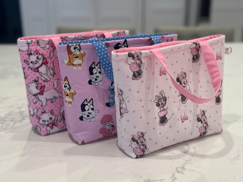 May include: Three fabric tote bags with different patterns. One bag features pink cats, another has Bluey characters, and the third showcases Minnie Mouse. Each bag has a pink handle and trim, and they are displayed on a white surface.