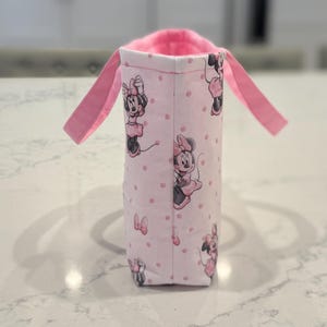 May include: A pink fabric bottle holder featuring a repeating pattern of Minnie Mouse. The holder has a pink interior and two pink handles. The exterior is light pink with small pink polka dots.