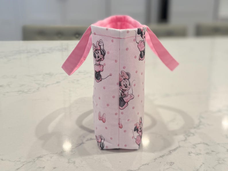 May include: A pink fabric bottle holder featuring a repeating pattern of Minnie Mouse. The holder has a pink interior and two pink handles. The exterior is light pink with small pink polka dots.