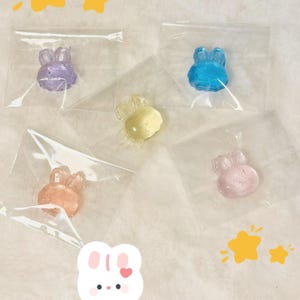 Puede incluir: Cinco pequeñas bolsas de plástico transparente que contienen cada una un caramelo con forma de conejo de un color diferente. Los caramelos son morado, azul, amarillo, rosa y naranja.