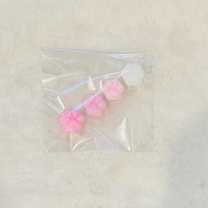 Könnte beinhalten: Ein klarer Plastikbeutel mit vier pfotenförmigen Wachsschmelzen. Drei sind rosa und einer ist weiß. Die Schmelzen sind in einer Reihe in dem Beutel angeordnet, der sich vor einem flauschigen, weißen Hintergrund befindet.