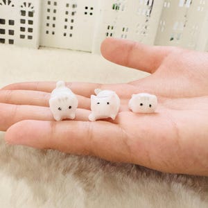 Pode incluir: Três pequenas estatuetas de cerâmica branca de gatos, cada uma com olhos pretos e uma pose diferente. As estatuetas estão sentadas na mão de uma pessoa.