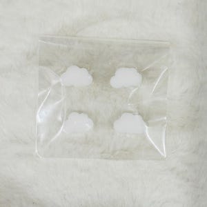 Puede incluir: Cuatro adornos blancos en forma de nube están encerrados en una bolsa de plástico transparente. Las nubes tienen una apariencia esponjosa. La bolsa está sobre una superficie blanca y texturizada.