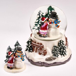 Puede incluir: Un globo de nieve con una escena de invierno con dos niños construyendo un muñeco de nieve. El globo está sobre una base con una valla de madera y árboles cubiertos de nieve. La base está decorada con un acabado de brillo plateado.