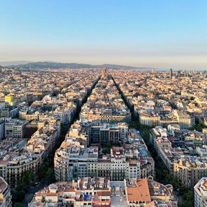 May include: An aerial view of a city with a grid pattern of streets lined with buildings. The buildings are mostly brown and beige, with some green trees and plants visible. The sky is a clear blue.