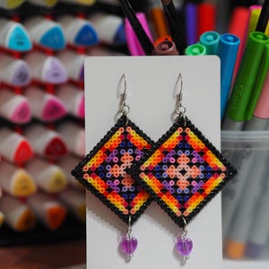 Könnte beinhalten: Ein Paar handgefertigte Ohrringe. Jeder Ohrring ist ein schwarzes Quadrat mit einem farbenfrohen geometrischen Muster in Orange, Gelb, Rosa und Lila. An jedem Ohrring hängt ein kleiner, lila Herzanhänger. Die Ohrringe werden auf einer weißen Karte präsentiert.