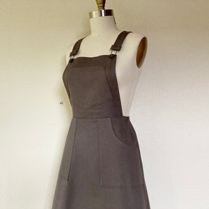 Peut inclure: Une robe salopette grise avec des bretelles réglables et une grande poche avant. La robe a deux poches supplémentaires à l'avant et est présentée sur un mannequin. La robe salopette est de forme trapèze et en tissu doux.