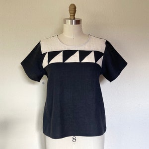 May include: Black and white short-sleeved top with a geometric design. The top features a black body with a white neckline and a horizontal band of alternating black and white triangles across the chest. The top is displayed on a mannequin.