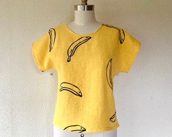 May include: A yellow short-sleeved top with a repeating black banana print. The top is displayed on a mannequin. The number "8" is printed in black at the bottom of the top.