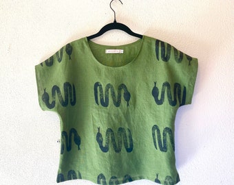 May include: Olive green linen top with a repeating black snake pattern. The short-sleeved shirt has a scoop neck and is hanging on a black hanger. The top is made of a lightweight fabric.
