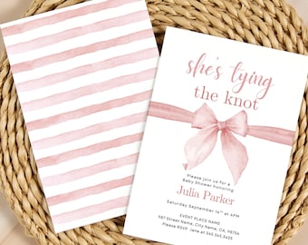 May include: Baby shower invitation with the text "she's tying the knot" in pink script. The invitation features a watercolor bow and ribbon design. A matching card with pink watercolor stripes is also present. The invitations are on a woven placemat.