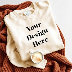 Könnte beinhalten: Ein cremefarbener Pullover mit dem Text "Your Design Here" in Schwarz auf der Vorderseite. Der Pullover ist gefaltet und auf einer braunen Strickdecke gelegt.