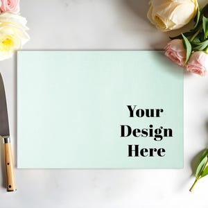 May include: A light green chopping board with the text "Your Design Here" on a white marble surface with pink and white flowers and a silver knife with a wooden handle.