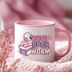 May include: A pink and white ceramic mug with a pink rim. The mug has a graphic of a stack of books, headphones, and the text "AUDIO BOOK WORM".