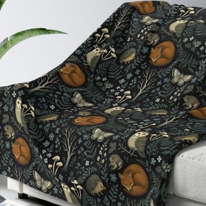 May include: A dark-coloured throw blanket featuring a repeating pattern of woodland creatures including foxes, owls, and hedgehogs, interspersed with illustrations of mushrooms, ferns, bare tree branches, and butterflies on a black background.