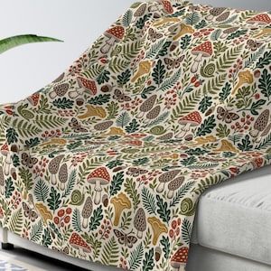 May include: A cream-coloured throw blanket featuring a repeating pattern of red and white mushrooms, yellow chanterelles, brown morels, green fern leaves, oak leaves, small red berries, acorns, snails, and brown moths scattered across the fabric.