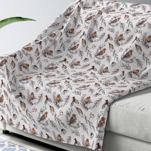 May include: A white throw blanket featuring a repeating pattern of small brown and orange birds perched among grey botanical illustrations of pine needles, holly leaves, and small berries, creating a detailed nature-inspired design.