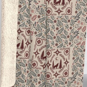 May include: A plush sherpa-lined throw blanket featuring a repeating pattern of dark red and black illustrations on a cream background. The design includes howling wolves, pine trees, crescent moons, stars, skulls, and various botanical leaf motifs.