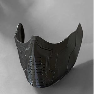 May include: A black plastic face mask with a geometric design. The mask has a textured surface and a raised pattern of lines and shapes.