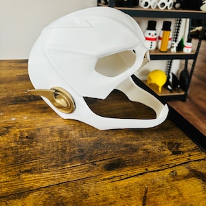 May include: White plastic The Flash helmet with a gold trim. The helmet is sitting on a wooden surface.