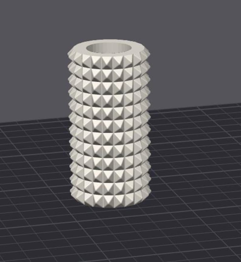 Op de afbeelding: Een cilindrisch, gebroken wit object met een reeks gestapelde, piramidevormige uitsteeksels. De bovenkant heeft een cirkelvormige opening. Het ontwerp is symmetrisch en geometrisch, waardoor een getextureerd oppervlak ontstaat.