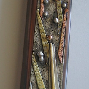 Puede incluir: Un colgante rectangular de metal con un marco de madera marrón. El colgante presenta un diseño de metal mixto con acentos dorados, plateados y cobrizos. El diseño incluye formas geométricas y una rama de árbol estilizada.