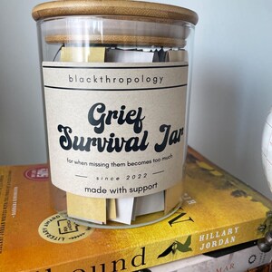 May include: A glass jar with a bamboo lid filled with small pieces of paper. The jar has a label that reads "Grief Survival Jar" with the text "for when missing them becomes too much since 2022 made with support".