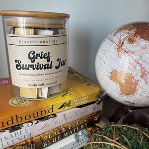 May include: A glass jar with a wooden lid filled with small pieces of paper. The jar has a label that reads "Grief Survival Jar" and "for when missing them becomes too much". The jar is sitting on a stack of books with a globe and a green moss ball.