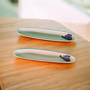 May include: Two oval-shaped, pale green glass serving dishes with a purple grape cluster design at one end. The dishes are displayed on a light-colored wooden surface, reflecting natural light.