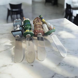 May include: Four stainless steel butter knives with decorative handles. The handles feature a coffee machine, a green and white coffee cup, a burlap sack, and a collection of condiments. The knives are arranged on a marble surface.