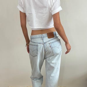 May include: A white short-sleeved cropped t-shirt with rolled sleeves paired with light-wash blue denim jeans. The jeans feature a brown leather patch on the back waistband, two back pockets with arcuate stitching, and a small red brand tab.